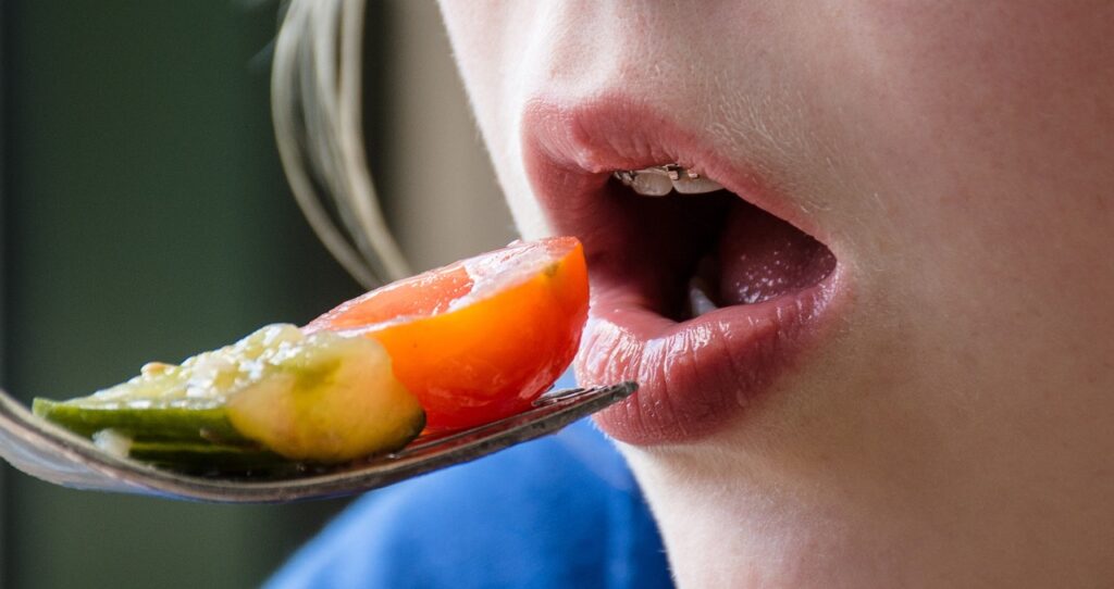 Nahaufnahme eines Mundes mit geöffneten Lippen, der eine Gabel mit Tomate und Gurke isst.